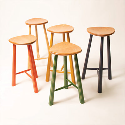 Five three-legged stools with triangular wooden seats and painted legs in various colors on a white background.