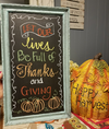 Rustic distressed wood chalkboard frame with colorful Thanksgiving-themed lettering and pumpkin drawings.