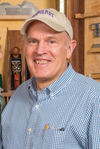 Man wearing a cap and checkered shirt standing in a woodworking workshop with hand planes on shelves behind him