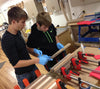 Two young men wearing gloves inspecting wood strips near bar clamps in a woodworking workshop