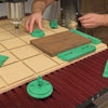 Hands positioning green clamps on a spoilboard grid with a wood blank on a CNC machine bed