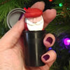 Hand holding a small carved wooden Santa head with red hat inside a black container against a Christmas tree background.