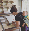 Woman marking measurements on plywood with a pencil and square while carrying a sleeping baby in a carrier in a woodworking