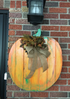 Wooden pallet slats assembled and painted orange as a pumpkin with burlap bow and foliage decoration hanging on brick wall