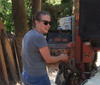 Woman adjusting controls on a woodworking machine outdoors, surrounded by wood and trees.