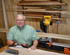 Woodworker in workshop with hand plane, miter saw, and stacked lumber behind him