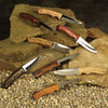 Seven custom knives with various wood handles displayed on stone slabs and gravel background