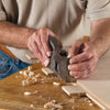Hands using a metal shoulder plane to trim a wooden tenon on a workbench with wood shavings.
